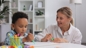 Adorable little african american boy with curly hair having fun at pediatric specialist appointment, happy child drawing with female therapist, exercises.