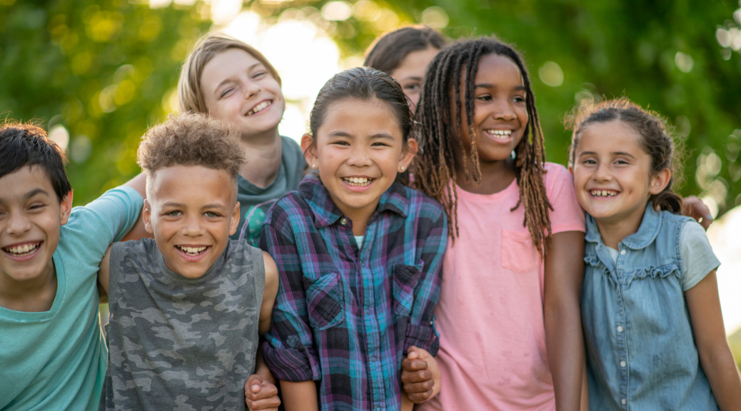 A large group of school aged children huddle together outside for a portrait. They are each dressed casually and are laughing and smiling as they enjoy each others company.