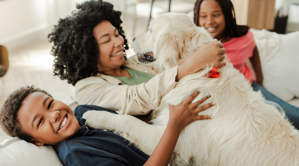 Happy family is relaxing on a sofa at home, embracing their golden retriever dog and smiling, therapy pets