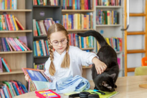 Elementary school girl reading a book and petting black cat at table.
